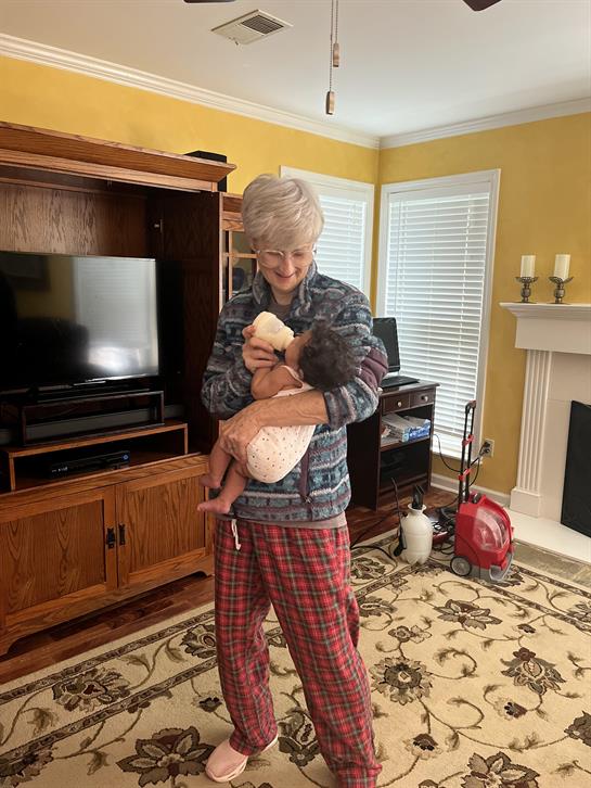 Elderly person feeding a baby with a bottle while standing in a warm and inviting living room.
