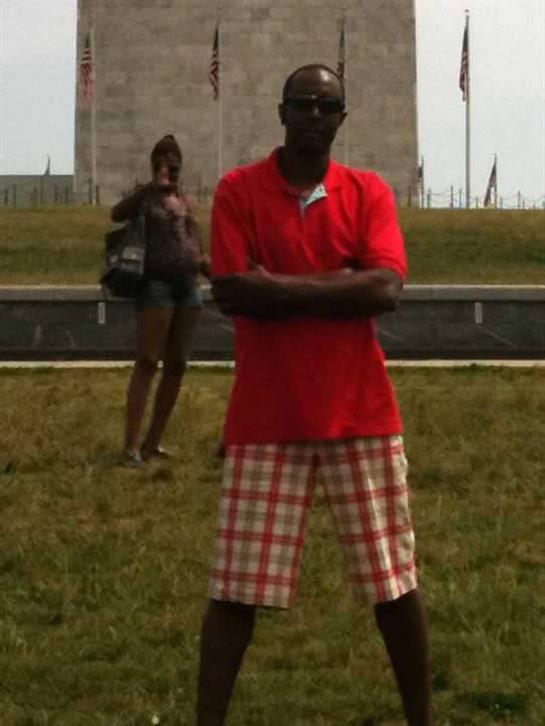 A man stands with arms crossed in a red shirt and plaid shorts while a woman poses behind him.