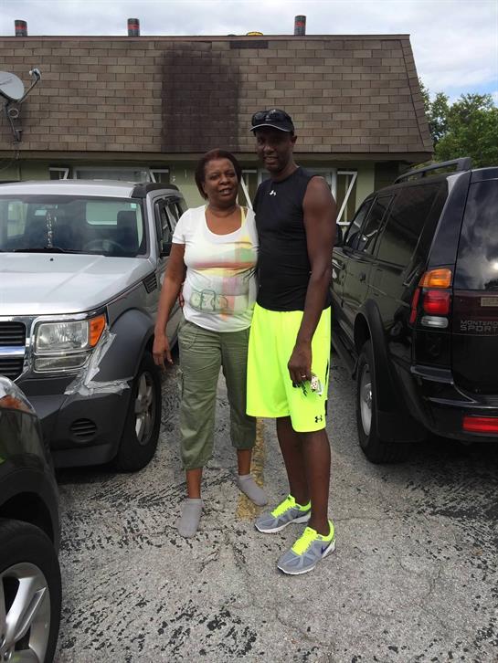 A couple smiles together in casual attire, surrounded by parked vehicles on a bright day.