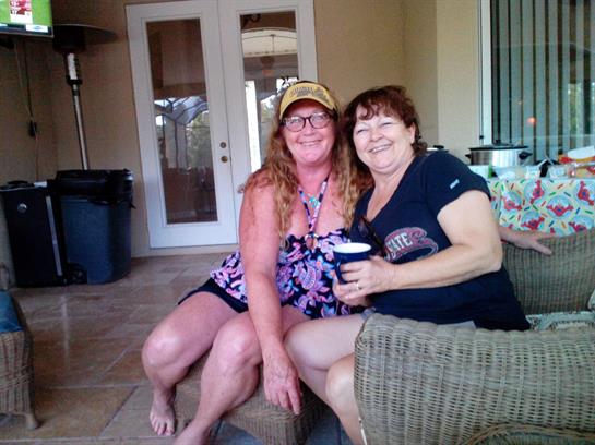 Two women sit on a patio, smiling and holding drinks, enjoying a sunny afternoon together.