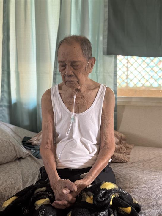 An elderly man sits on a bed wearing an oxygen tube, lost in thought in a sunny room.