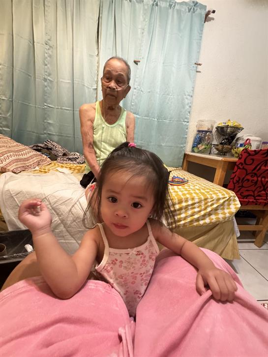 A young girl plays in the foreground while her grandmother watches from the bed in a warm space.