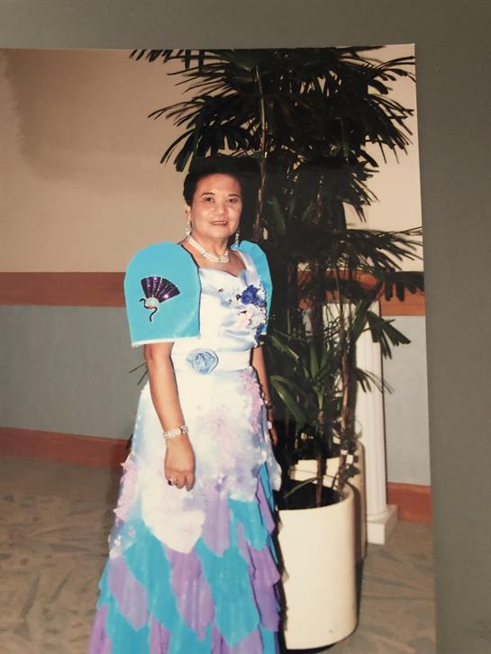 A woman dressed in a vibrant gown stands proudly next to a decorative plant indoors.