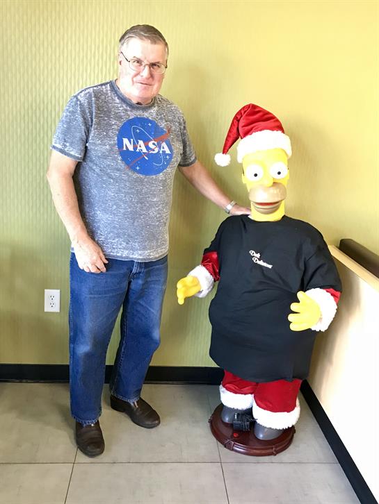 An adult man stands next to a festive cartoon character dressed in Santa attire, smiling.