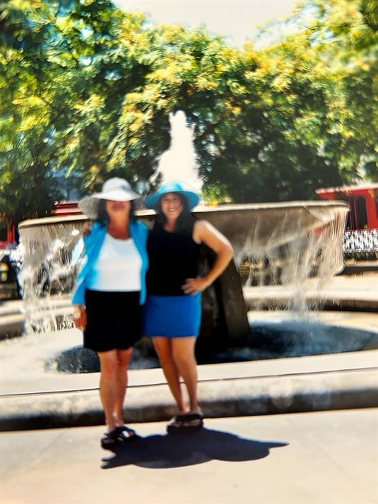 Two women stand near a fountain, enjoying the warm weather in a lively park.
