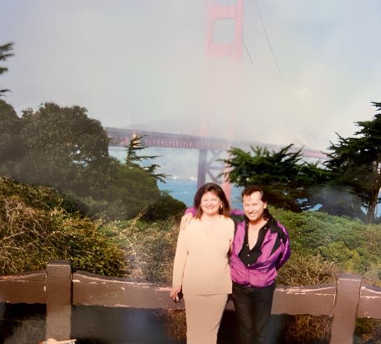 A couple stands by a wooden railing smiling, with the Golden Gate Bridge partially visible in fog.