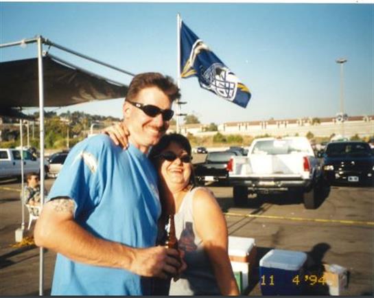 A couple smiles together while holding drinks at an outdoor gathering under clear skies.