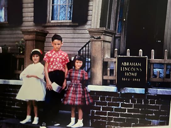 Three children in vintage clothing pose joyfully outside a historic house on a sunny day.