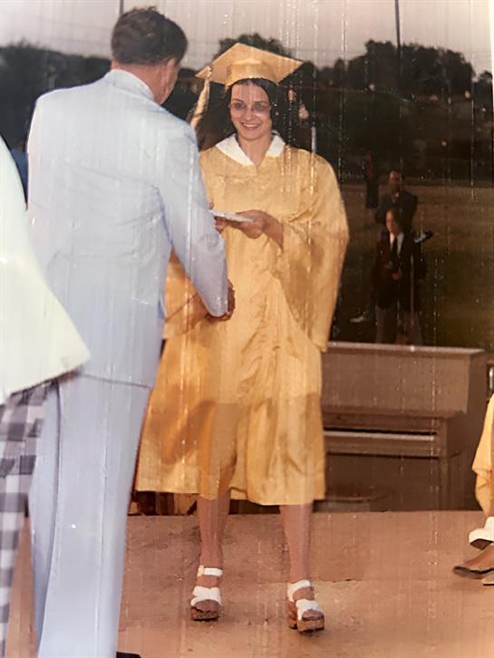 A student in a yellow graduation gown proudly accepts a diploma during a ceremony held outdoors.