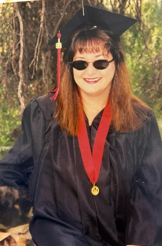 A proud graduate wearing sunglasses and a black gown outside with a smile on her face.