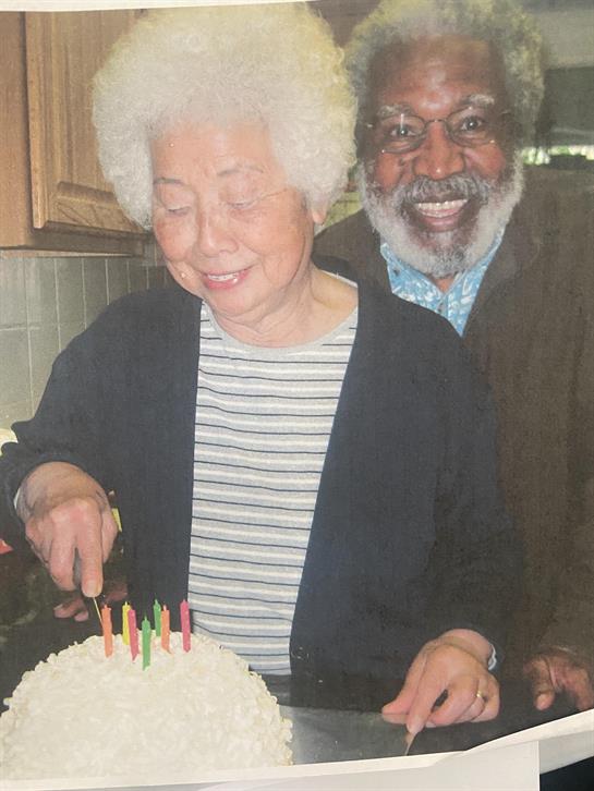 A cheerful elderly woman cuts a candlelit cake as a smiling man watches in the kitchen.