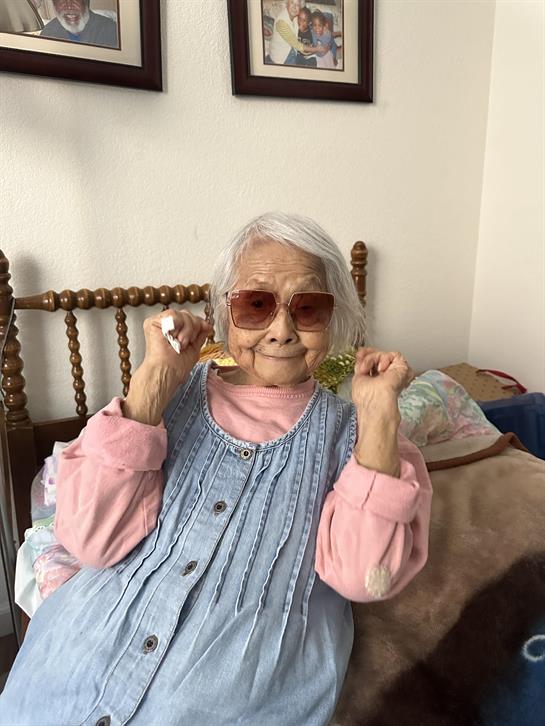 An elderly woman enjoys her time at home, wearing sunglasses and playful earrings while sitting.