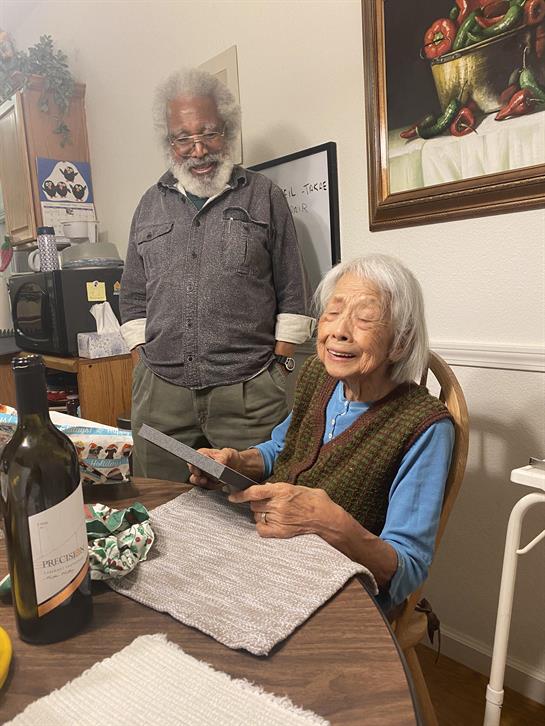An elderly woman joyfully looks through a photo album while a man watches her at home.