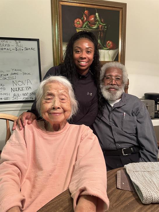 A young woman poses with her grandparents while smiling warmly in their home together.