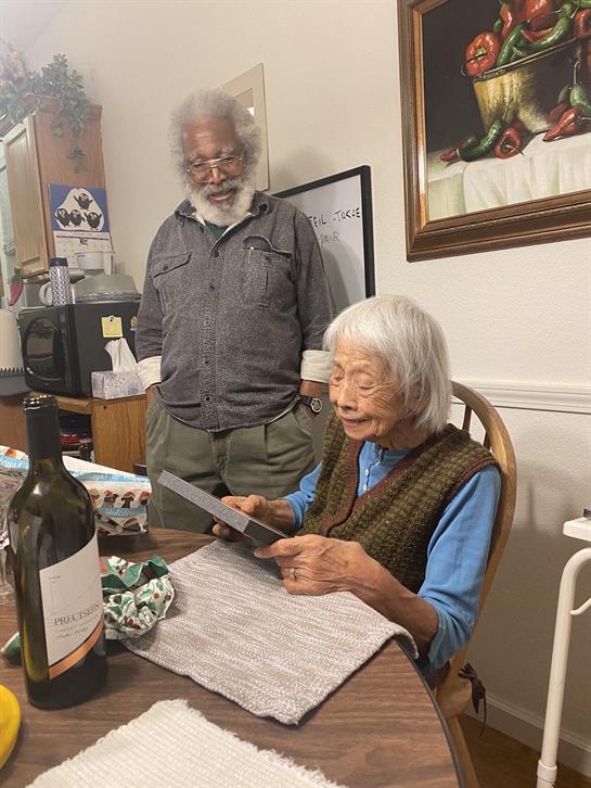An elderly woman examines a letter at the dining table. A man stands nearby, watching her closely.