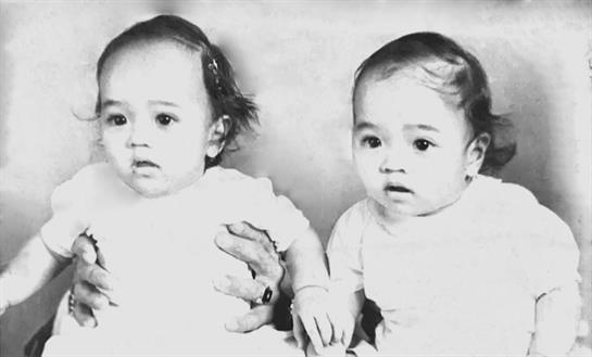 Two identical twins are seated side by side, gazing at the camera while being held.