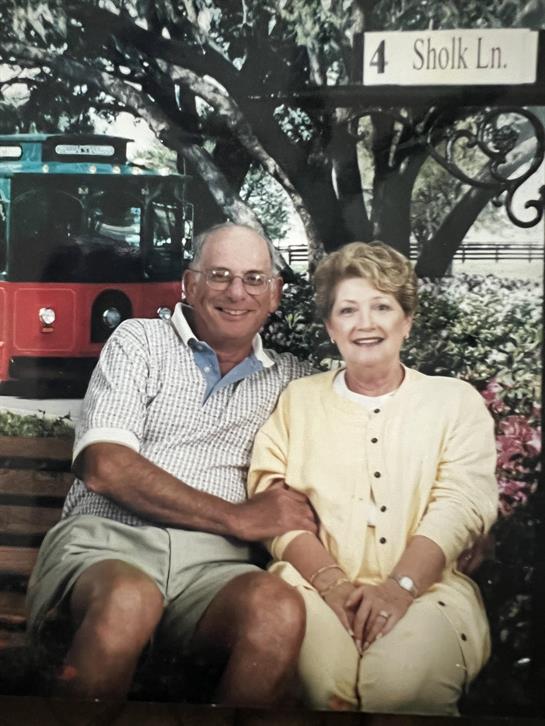 Elderly couple smiles while sitting on a park bench, surrounded by greenery and a vintage trolley.