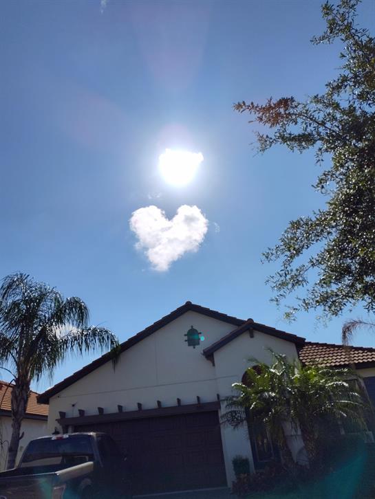 A heart-shaped cloud is visible in the clear sky above a suburban house, with sunshine.