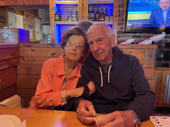 An elderly couple shares a warm moment at a wooden table in a rustic restaurant setting.