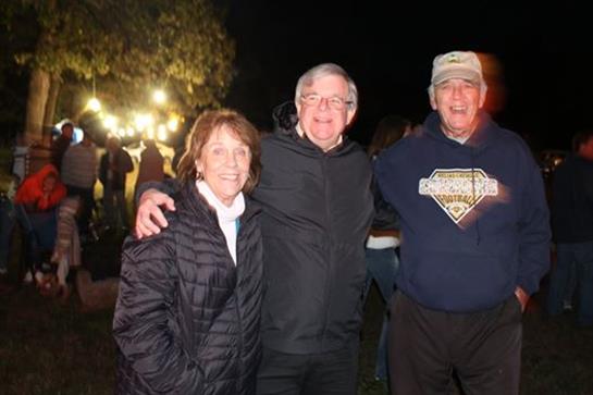 Three friends pose together outdoors at a nighttime community event with lights and people around.