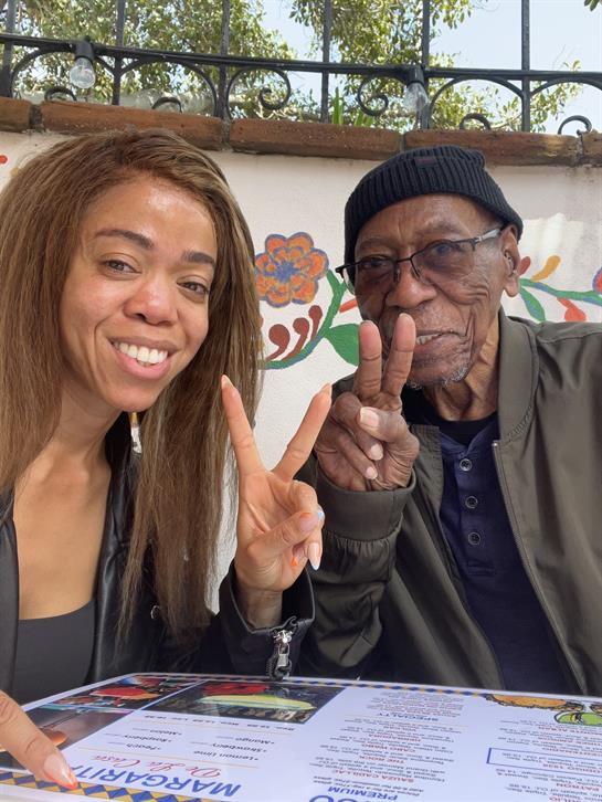 Two individuals are happily posing together, making peace signs at a cafe table.