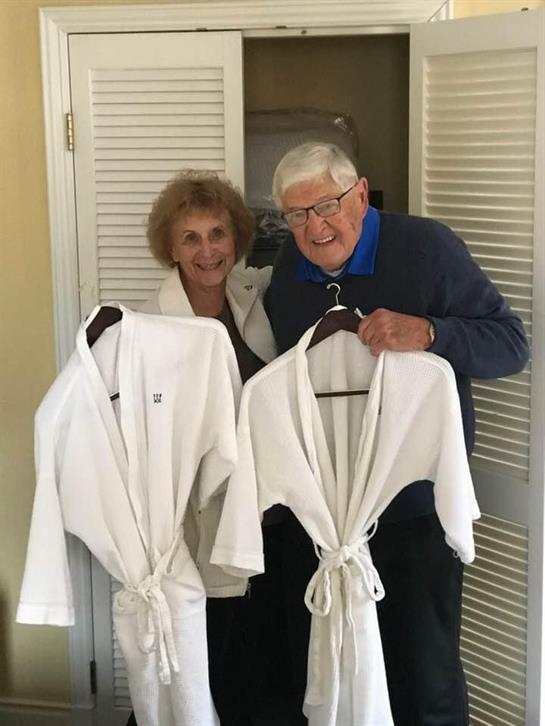 An elderly couple smiles brightly while holding matching white bathrobes in a warm setting.
