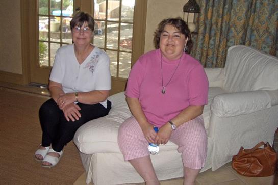 Two women chat while sitting in a bright living room with sunlight streaming in.