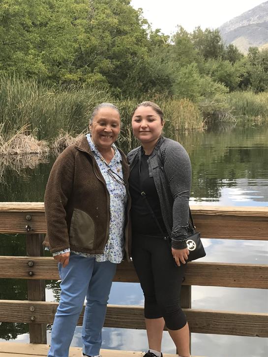 Two women smile as they stand on a bridge overlooking a tranquil lake with lush plants.