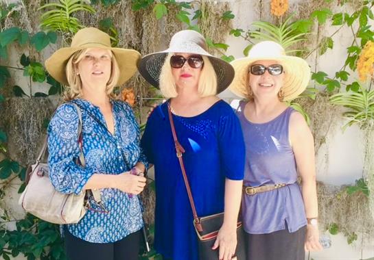 Friends pose in a lush garden, sporting vibrant outfits and sun hats on a sunny day.