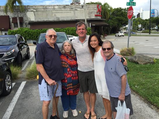 Five friends pose happily together on a sunny day near a parking lot.