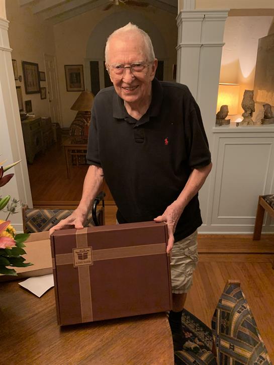 An elderly man stands in a warm, inviting room, joyfully presenting a brown gift box.