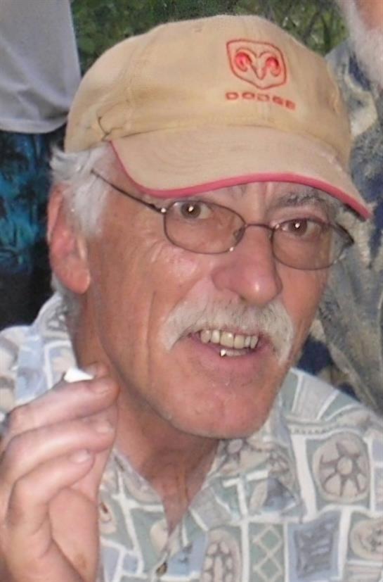 A joyful man sits outdoors wearing glasses and a cap, chatting with friends and holding a cigarette.