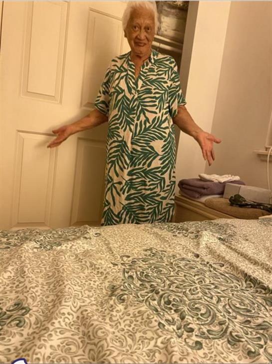 An elderly woman in a floral dress stands with arms outstretched near a closed door.