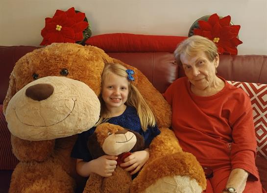 A young girl hugs a large teddy bear while sitting with her grandmother, embracing joy.