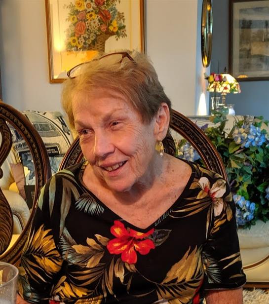 An elderly woman with glasses smiles warmly as she enjoys a drink during a relaxed gathering.