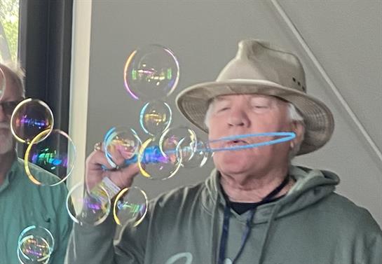 An elderly man skillfully blows bubbles during a joyful indoor gathering with friends around.
