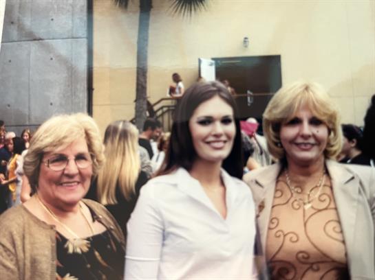 Three women are smiling together at a lively event with a bustling background full of attendees.