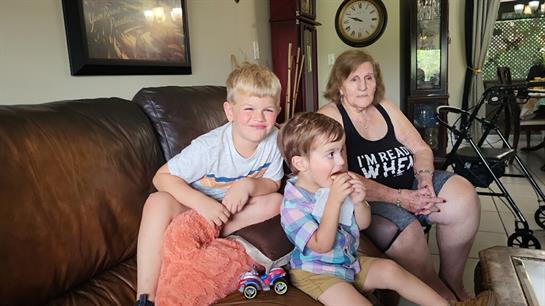 Two young boys are seated on a couch while a woman watches them in a cozy living room.