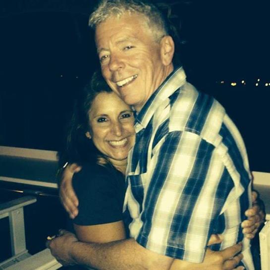 Two people share a warm hug while smiling in a cozy outdoor setting at night.