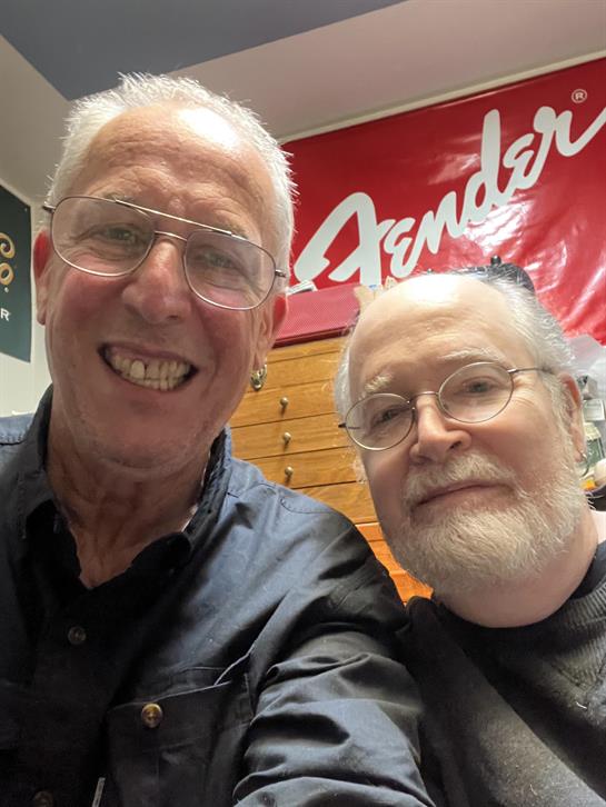 Two men pose for a cheerful selfie in a music studio surrounded by guitars.