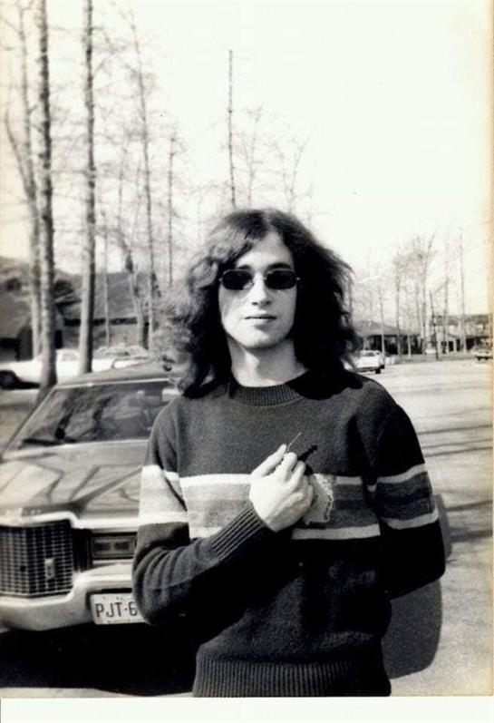 A young man with long hair and sunglasses poses beside a parked car under bare trees.