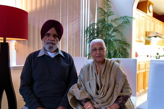 Elderly couple sits comfortably in a bright living area with plants and stylish furnishings.