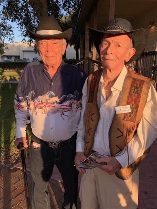Two men dressed in cowboy outfits stand together, enjoying an outdoor gathering in the evening.