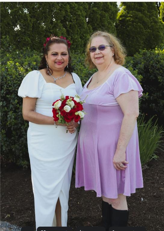 Two women pose in a garden, one in a white dress and the other in purple, surrounded by flowers.