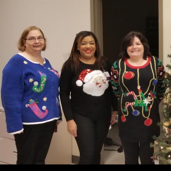 Three women in colorful holiday sweaters pose joyfully in an office decorated for Christmas.