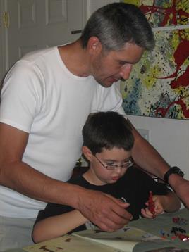 A father helps his son assemble colorful building blocks at a table in their home.