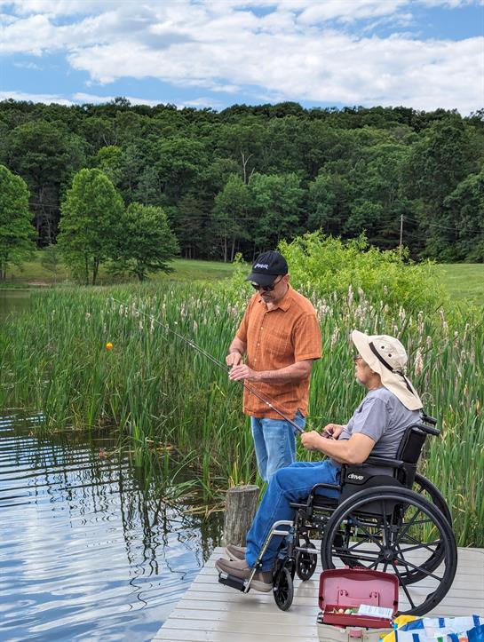 A man helps a woman in a wheelchair learn to fish beside a calm lake surrounded by greenery.