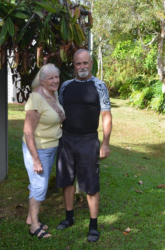 Elderly couple stands together smiling amidst greenery, enjoying a sunny day in the garden.