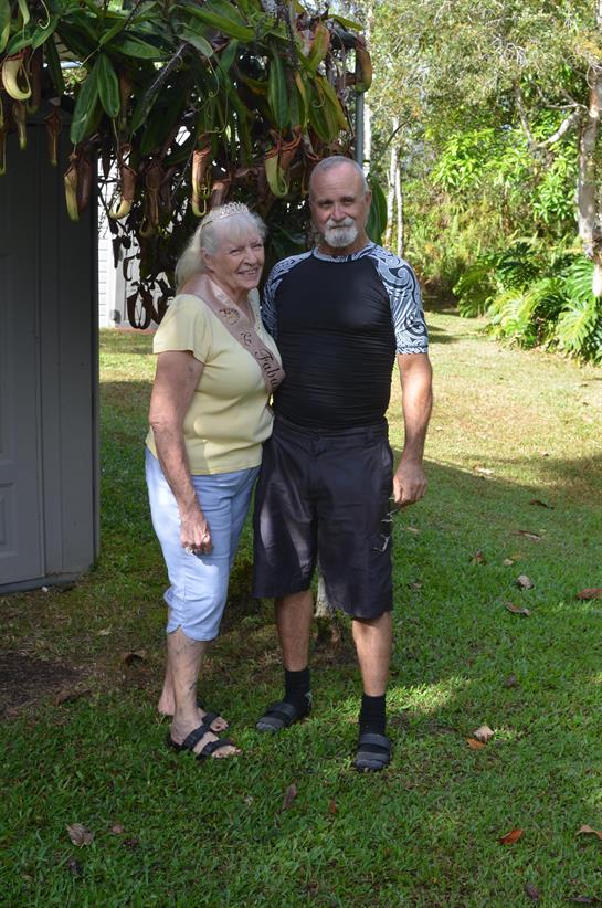 An elderly couple smiles while standing in a vibrant garden filled with greenery.