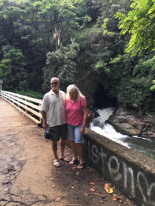 A couple stands by a river, surrounded by green trees and a tranquil waterfall nearby.